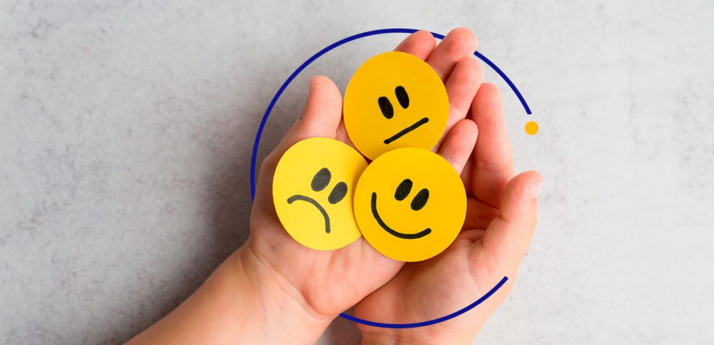 Mãos segurando 3 recortes de carinhos parecidos com emojis de felicidade e tristeza. Esse conjunto simboliza inteligência emocional.